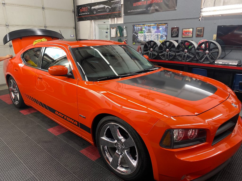 Orange Dodge Charger Daytona with black hood detail parked in a garage; trunk open, custom wheels displayed on wall.
