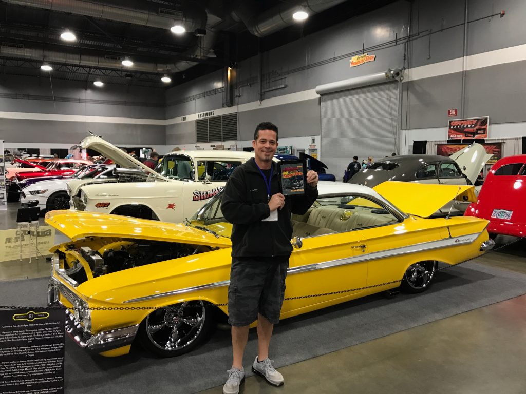 Man stands next to a yellow vintage car with its hood open, holding a plaque at an indoor car show.