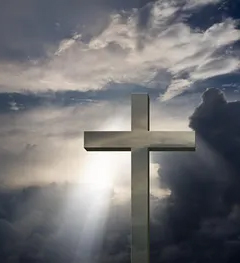 A cross stands against a cloudy sky with sunlight streaming through the clouds in the background.