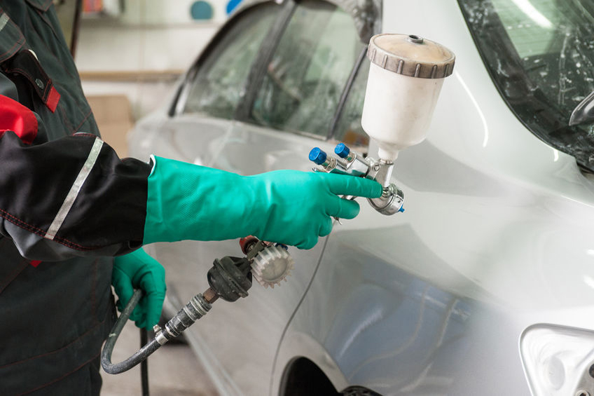 Person wearing green gloves uses a spray gun to paint the side of a silver car in an auto body shop.