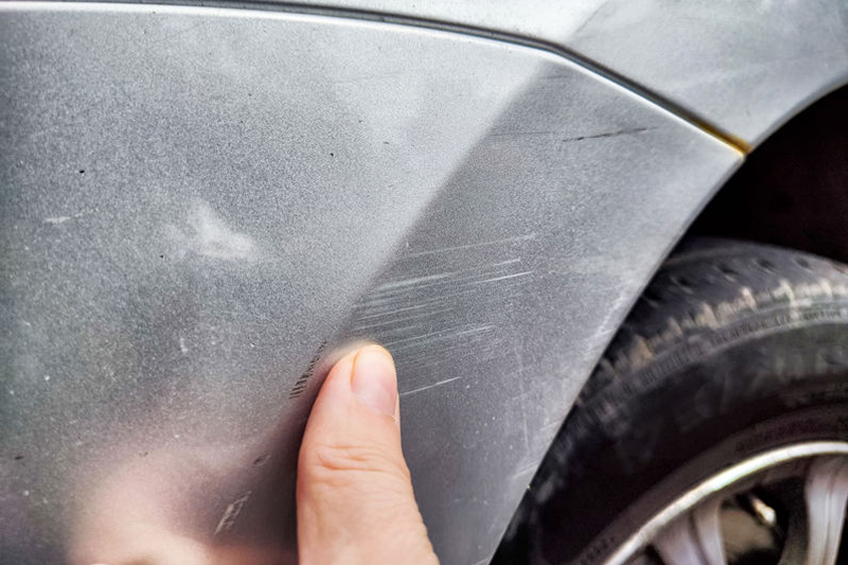 Close-up of a hand touching light scratches on the silver paint of a car near the wheel.