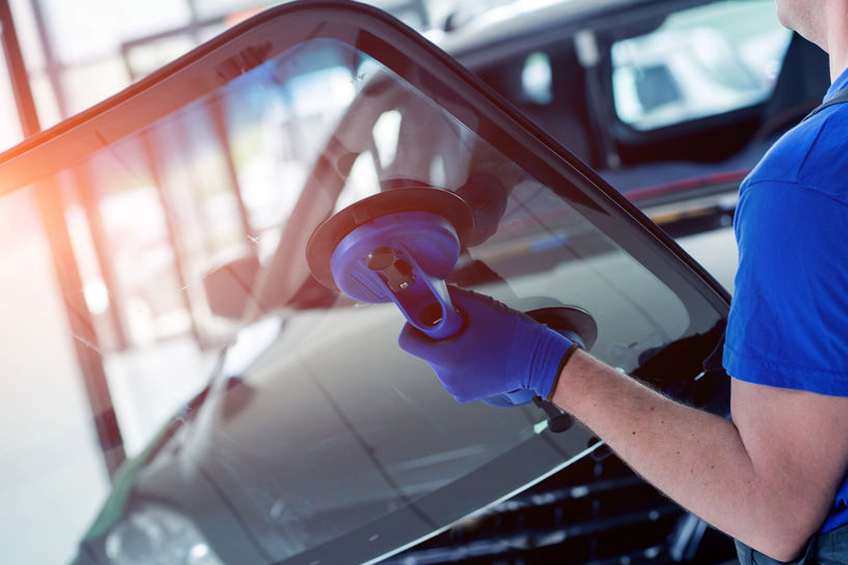 Person wearing blue gloves uses a suction tool to install or remove a windshield from a car in a workshop.
